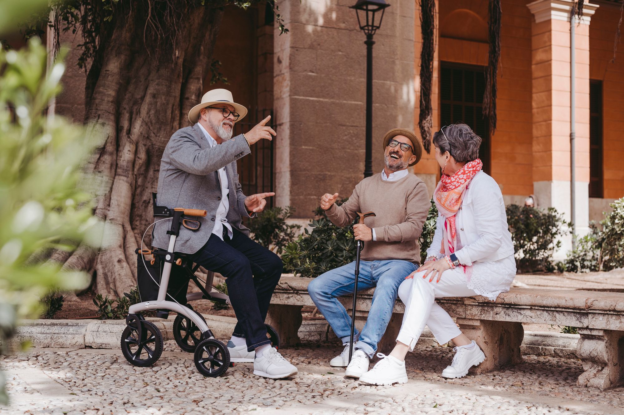 Drei ältere Menschen lachen miteinander auf einer Parkbank. Ein älterer Herr sitzt auf einem Rollator.